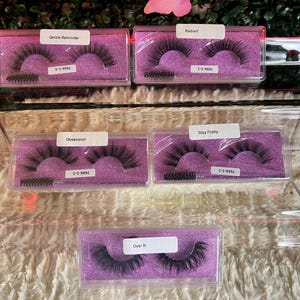 K&ouml;nnte beinhalten: F&uuml;nf Paar falsche Wimpern in durchsichtiger Plastikverpackung mit violetten Deckeln. Die Wimpern sind mit den Namen "Gentle Reminder", "Radiant", "Obsession", "Stay Pretty" und "Over It" beschriftet.