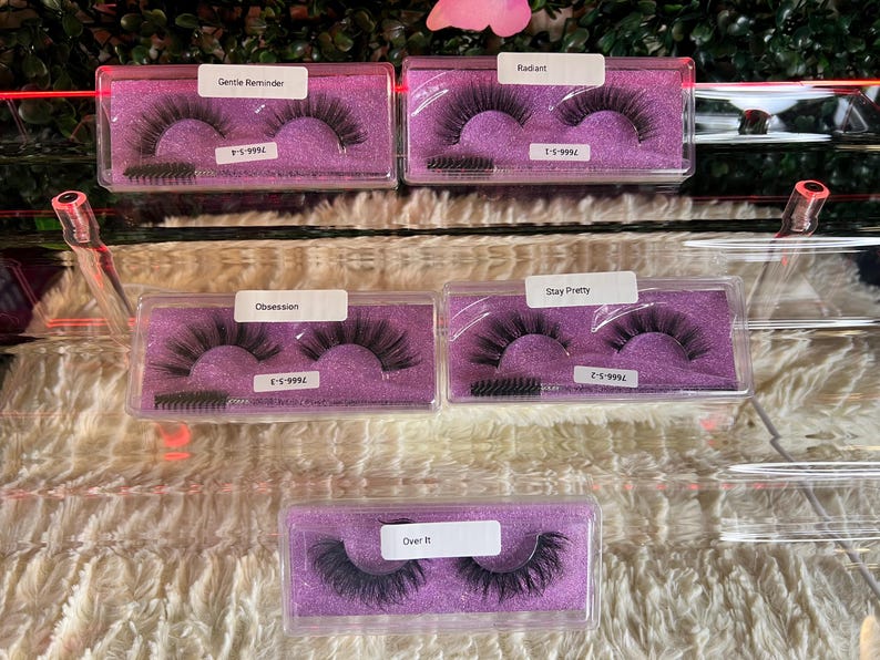 K&ouml;nnte beinhalten: F&uuml;nf Paar falsche Wimpern in durchsichtiger Plastikverpackung mit violetten Deckeln. Die Wimpern sind mit den Namen "Gentle Reminder", "Radiant", "Obsession", "Stay Pretty" und "Over It" beschriftet.