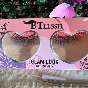 K&ouml;nnte beinhalten: Eine rosa und violette Schachtel mit einem herzf&ouml;rmigen Fenster, in dem ein Paar k&uuml;nstliche Wimpern zu sehen ist. Auf der Schachtel steht "BTLLSSH" und "GLAM LOOK NATURAL HAIR".