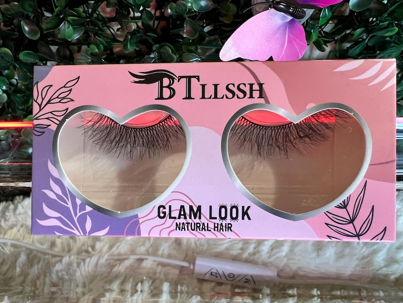 K&ouml;nnte beinhalten: Eine rosa und violette Schachtel mit einem herzf&ouml;rmigen Fenster, in dem ein Paar k&uuml;nstliche Wimpern zu sehen ist. Auf der Schachtel steht "BTLLSSH" und "GLAM LOOK NATURAL HAIR".