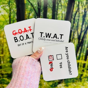 May include: Three white square coasters with humorous text. One reads "G.O.A.T B.O.A.T (BIT OF A TWAT)", another says "T.W.A.T Totally wise and talented", and the third asks "Are you childish?" with a "Knob" option.