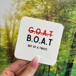 Può includere: Un adesivo quadrato bianco con la scritta "G.O.A.T" sbarrata e sostituita da "B.O.A.T (BIT OF A TWAT)" in nero e rosso. L'adesivo è tenuto davanti a uno sfondo verde e giallo sfocato.