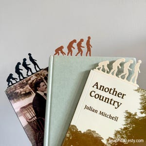 May include: Three evolution-themed bookmarks in black, brown, and cream colors, placed on top of books. The bookmarks depict the evolution of humans. One bookmark is on a book titled "Another Country" by Julian Mitchell.
