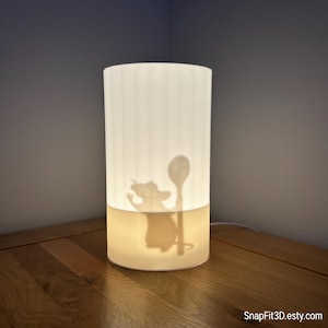 May include: A cylindrical lamp with a white, ribbed shade. The lower section glows with a warm light, displaying a silhouette of a cartoon rat holding a spoon. The lamp sits on a wooden surface. The Etsy shop name "SnapFit3D.etsy.com" is visible.