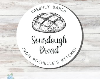 May include: A circular white sticker featuring a black ink illustration of a loaf of bread at the top. The text reads Freshly Baked, Sourdough Bread, and From Rochelle's Kitchen in a mix of sans-serif and script fonts arranged around the bread.
