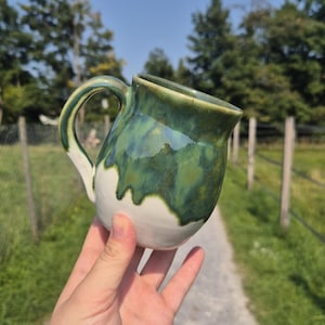 Könnte beinhalten: Handgefertigte Keramiktasse mit einzigartigem Design. Die Tasse hat einen weißen Boden mit welligem Rand, der in eine grüne und blaue Glasur übergeht. Der Griff ist grün. Perfekt für Kaffee oder Tee.