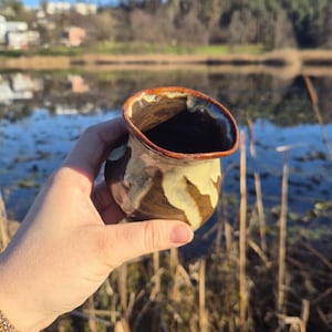 May include: A small, hand-thrown ceramic cup with a brown, white and orange glaze. The cup has a rounded shape and a slightly flared rim.