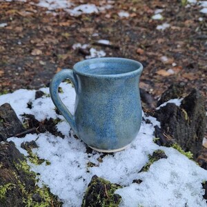 May include: A blue ceramic mug with a speckled glaze. The mug is sitting on a tree stump covered in snow.