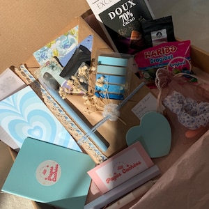 Peut inclure: Une boîte cadeau contenant divers articles, dont une tablette de chocolat Lindt Excellence 70% Cacao, des bonbons Haribo Dragibus, des articles de papeterie, des stylos et un carnet bleu. La boîte comprend également des éléments décoratifs et un chouchou.