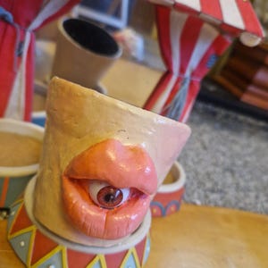 May include: A quirky ceramic sculpture of a face with prominent pink lips and a single, realistic eye. The sculpture is set against a backdrop of a circus tent with red and white stripes, and is decorated with colourful geometric patterns.