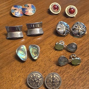 May include: A collection of six pairs of earrings featuring various designs including round sand dollar motifs, polished silver-tone bars, iridescent abalone shell shapes, red cabochon stones, dangling green faceted gems, and circular textured metal discs.
