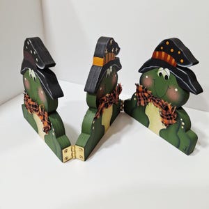 May include: A three-paneled wooden screen featuring green frog figures wearing black hats with orange and black plaid accents. Each frog has a matching plaid scarf around its neck. The panels are connected by gold-colored metal hinges.