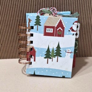 May include: A small spiral-bound notebook with a light blue cover featuring a Christmas-themed pattern of green pine trees, a red house labeled SANTA, a snowman, and a reindeer. The binding consists of copper-colored metal wire coils.