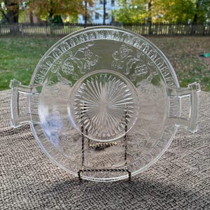 May include: Clear glass serving tray with etched floral and geometric patterns. The tray has a circular design with a central starburst pattern and two rectangular handles. It is displayed on a decorative metal stand.