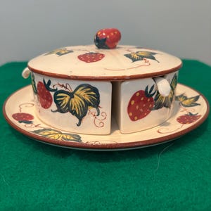 May include: A ceramic strawberry-themed serving dish set. The set includes a round plate, a lidded bowl, and two divided compartments. The bowl and plate are decorated with red strawberries and green leaves on a cream background. The lid has a strawberry-shaped knob.