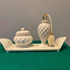 May include: A white ceramic vanity set featuring a lidded jar, a perfume bottle with a bulb atomizer, and a tray. The set has a quilted diamond pattern. The tray has wing-like sides.