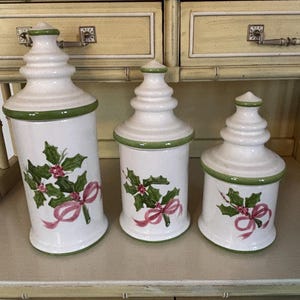 May include: Set of three white ceramic canisters with green trim and lids. Each canister features a holly and berry design with a pink ribbon. The canisters are different sizes and are arranged on a light-colored shelf.