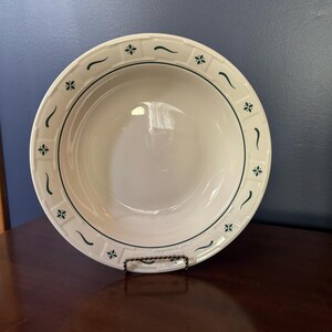 May include: A white ceramic bowl with a green floral and curved line pattern around the rim. The bowl is displayed on a dark brown surface against a blue background. The bowl is approximately 10 inches in diameter.