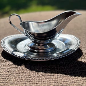 May include: A silver gravy boat with a curved handle and a pouring spout, resting on a matching round plate. The plate has a decorative beaded edge. The items are made of shiny metal and are set against a neutral background.