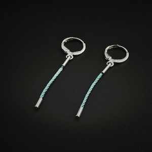 Könnte beinhalten: Ein Paar silberfarbene Creolen mit hellblauem, gedrehtem Kordeldesign. Die Ohrringe haben einen schlichten, eleganten Stil mit einem kleinen silbernen Zylinder am unteren Ende der Kordel. Der Hintergrund ist schwarz.