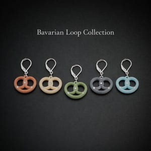 Könnte beinhalten: Fünf Paar Ohrringe in Brezelform in den Farben Orange, Beige, Grün, Grau und Blau. Jeder Ohrring hat einen silberfarbenen Haken und ist mit kleinen, klaren Kristallen verziert. Der Text "Bavarian Loop Collection" steht über den Ohrringen.