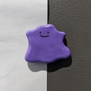 Könnte beinhalten: Ein lila, tropfenförmiger Magnet mit einem lächelnden Gesicht. Der Magnet hat eine einfarbige Farbe und eine glatte Textur. Der Hintergrund ist zwischen Weiß und Grau geteilt.