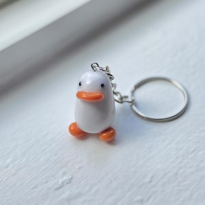 Puede incluir: Un llavero de pato blanco con un cuerpo liso y redondeado, un pico de color naranja brillante y dos pequeñas patas naranjas. El pato tiene dos diminutos ojos negros y está unido a una anilla de metal plateado mediante una cadena corta.