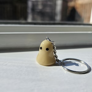 May include: A small, cream-coloured ghost keyring with black eyes. The ghost is attached to a silver key ring.