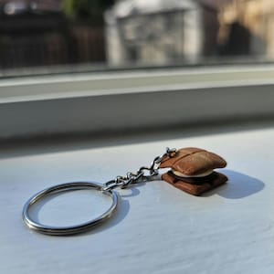 May include: A silver keyring with a small, brown and white s'mores charm. The charm is designed to resemble a graham cracker sandwich with a marshmallow and chocolate filling.