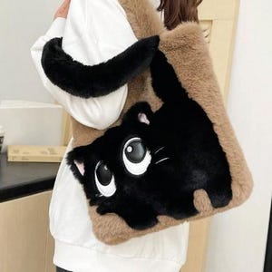 Könnte beinhalten: Eine flauschige Tragetasche mit einem Cartoon-Katzen-Design. Die Tasche hat eine schwarze Katze mit großen weißen Augen und eine flauschige braune und schwarze Außenseite. Der Riemen ist schwarz und die Tasche ist rechteckig.