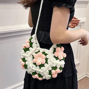 Puede incluir: Un bolso de crochet blanco con un diseño floral. El bolso presenta una serie de cuadrados blancos con flores rosas y hojas verdes. El bolso tiene una correa trenzada blanca.