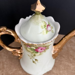 May include: A white porcelain teapot with a gold trim and floral design. The lid has a gold finial and a floral design. The teapot is decorated with pink roses and green leaves.