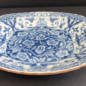 May include: A square ceramic dish with rounded corners featuring a blue and white floral pattern. The center displays a cluster of stylized flowers and leaves, while the border is decorated with a cross-hatched lattice design and foliage.