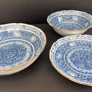 May include: A set of three ceramic dishes featuring a white base with intricate blue floral and vine patterns. The collection includes one large shallow bowl, one smaller deep bowl, and one flat plate, all finished with a thin gold-toned rim.