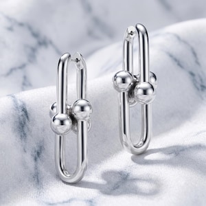 May include: A pair of silver-toned hoop earrings featuring an elongated oval link design. Each earring is accented with three polished spherical beads clustered at the center of the vertical bar, creating a structured and reflective metallic appearance.