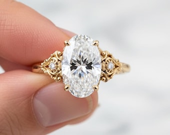 May include: A gold engagement ring featuring an oval-cut diamond. The ring has intricate filigree detailing on the band and around the diamond, with smaller accent stones. The ring is held up by a person's fingers.