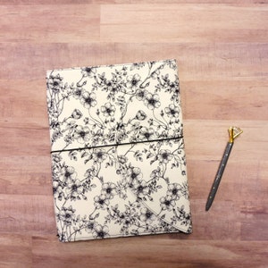 May include: A white fabric journal with a black floral pattern. The journal has a black elastic closure and is lying on a wood surface.