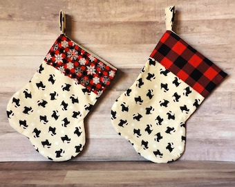 May include: Two Christmas stockings with a cream background and black dog silhouettes. One stocking has a red and black plaid cuff, the other has a red cuff with white snowflakes. Both have a cream loop for hanging.