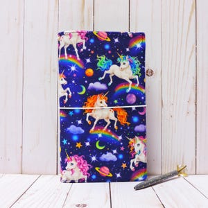 May include: A dark blue notebook cover with a colorful unicorn and rainbow pattern. The cover features white stars, moons, and planets on a dark blue background.