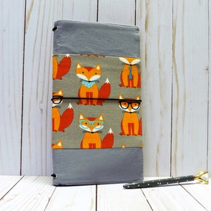 May include: A gray fabric journal cover with a pattern of orange foxes wearing glasses and ties. The cover has a black elastic band to hold it closed.