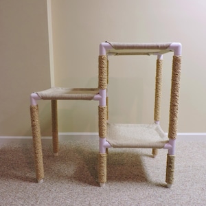 May include: A two-tier cat tree made of white PVC pipe wrapped in natural sisal rope. The cat tree has two platforms covered in beige fabric.