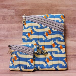 May include: A set of three quilted fabric bags with a dachshund dog pattern. The bags feature a blue and white striped background with repeating images of dachshunds. Each bag has a gray zipper and a silver-colored metal zipper pull.