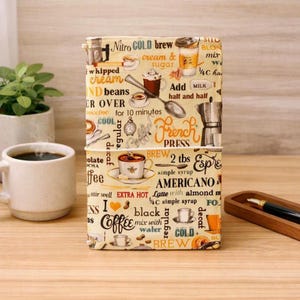 Può includere: Una copertina per taccuino rettangolare con sfondo color crema e una stampa ripetuta a tema caffè. Il design include illustrazioni di tazze da caffè, una caffettiera a stantuffo, cucchiai e testi come Nitro cold brew, Americano e Coffee.