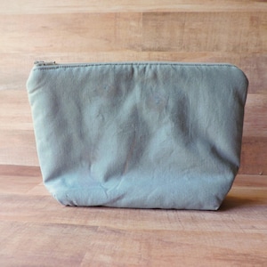 Può includere: Una pochette con cerniera di colore grigio chiaro realizzata in tessuto. La pochette è appoggiata su una superficie di legno.