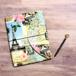 May include: A vintage-style journal with a colorful fabric cover featuring an image of the Eiffel Tower, flowers, and handwritten text. The journal is closed and has a black elastic band around it. A gold-colored pen is lying next to the journal.