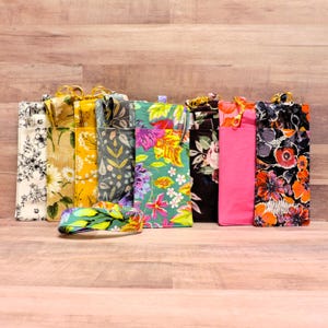 May include: A collection of eight rectangular fabric pouches with gold-toned metal clips, featuring various floral and botanical patterns in colors including white, yellow, gray, green, pink, and black, with one matching strap lying in front.