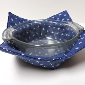 Puede incluir: Un calientaplatos de tela azul y blanco con un cuenco de vidrio en su interior. El calientaplatos está hecho de tela de algodón y tiene un patrón geométrico de círculos y cruces blancos sobre un fondo azul.