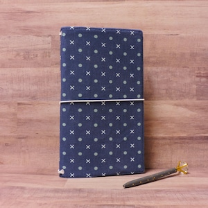Puede incluir: Una cubierta de cuaderno de tela azul con un patrón geométrico blanco y verde. La cubierta tiene una banda elástica blanca.