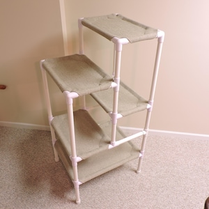May include: A multi-level cat tree made of white PVC pipe with beige fabric covered platforms. The cat tree has four levels and is freestanding.