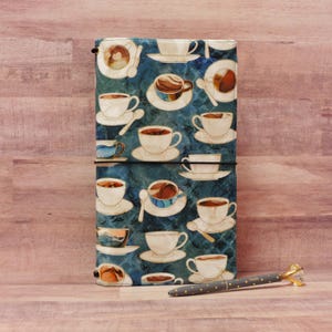 May include: A blue and white fabric notebook cover with a pattern of coffee cups and saucers. The cover has a black elastic band and a black pen with gold accents is lying on a wooden surface.